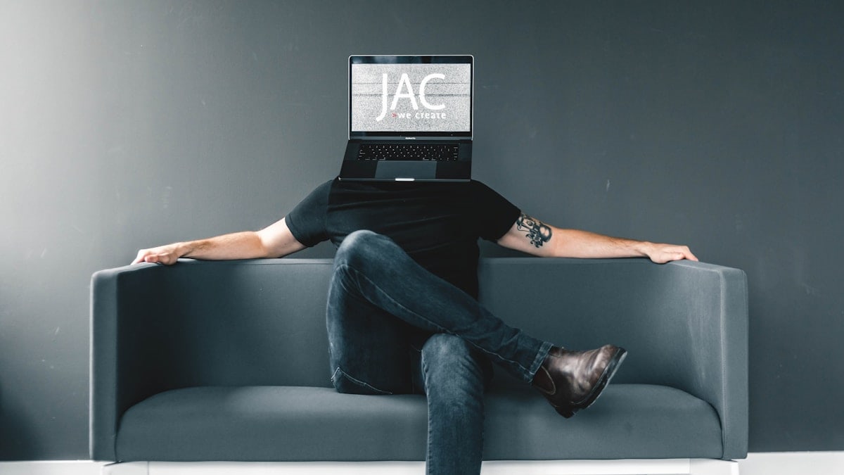 man in black shirt and blue jeans sitting on gray sofa with JAC mask